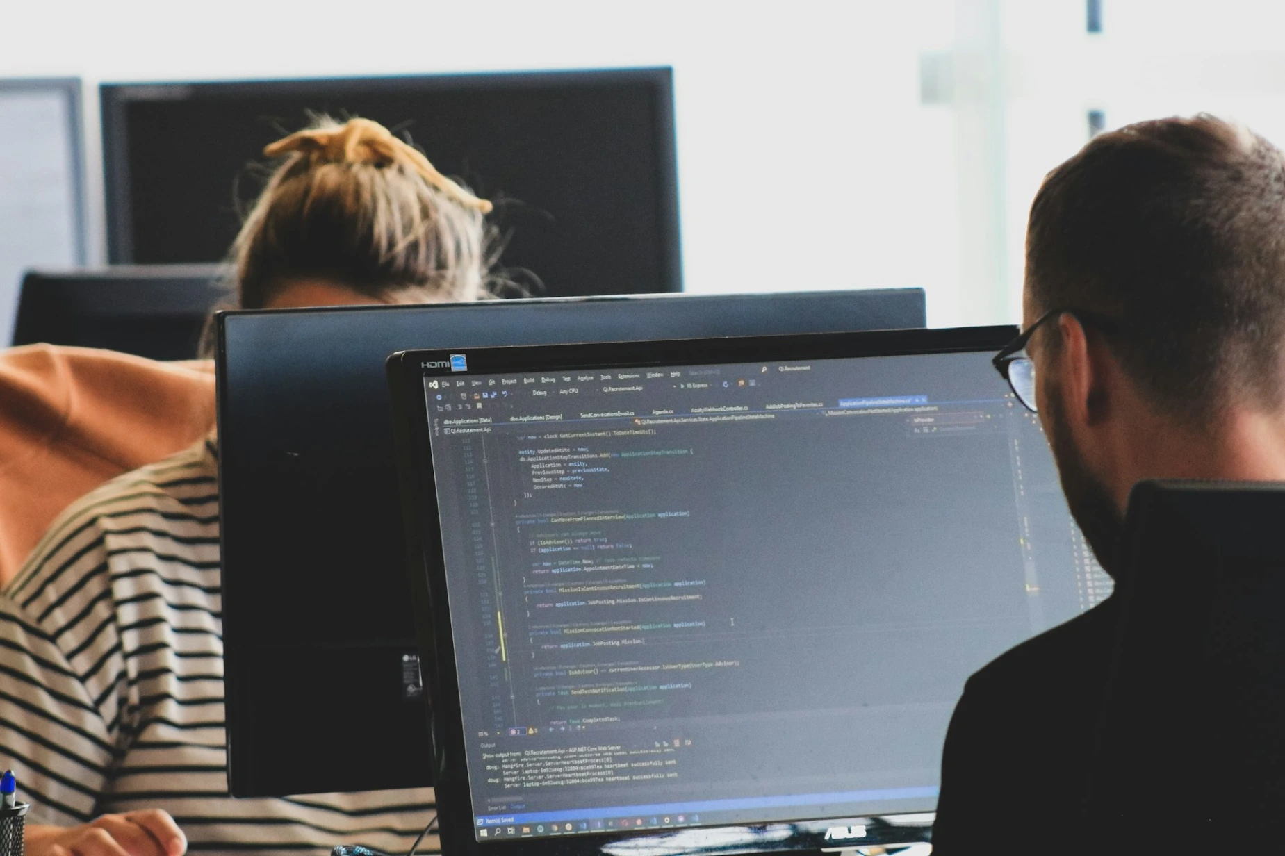 Two people working on computer code at monitors in a bright office workspace